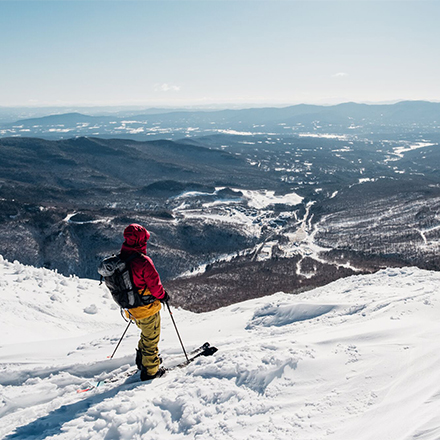 A person on skis at an overlook taking in a view of mountains and towns on a sunny winter day.&nbsp;