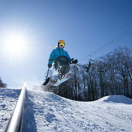 A person using adaptive skiing equipment hits a jump on a sunny winter day on a ski slope.&nbsp;