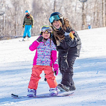  A child and an adult on snowboards smiling at the camera on a sunny winter day. 