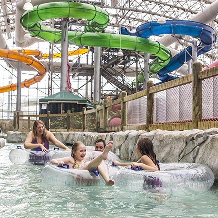 A family plays in a lazy river in an indoor waterpark, laughing and swimming toward the camera.