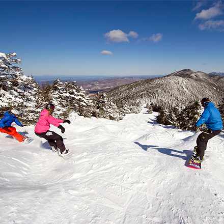 Three people snowboard down a mountain on a sunny day with sweeping mountain views visible.&nbsp;