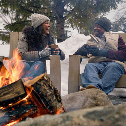 Two people sit by a fire with drinks, talking and laughing, outdoors in the winter.