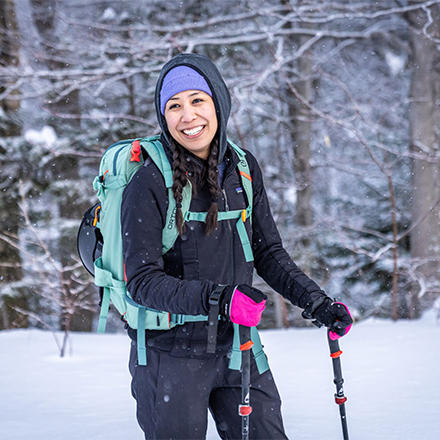 A person wearing a backpack on skis smiling at the camera outside on a snowy trail