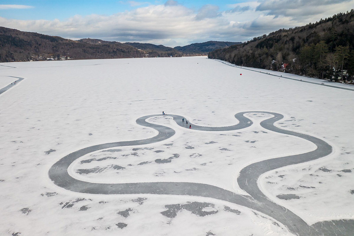 Seen from above, a winding trail on a frozen lake with skaters. Mountains are visible in the background on a winter day. 
