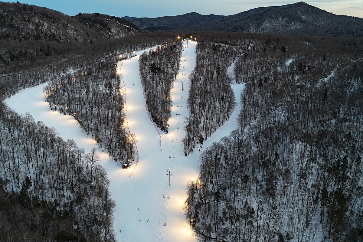 Seen from far above in an aerial shot, a ski area with chairlifts and lighting at dusk in the winter with snow covering trails.