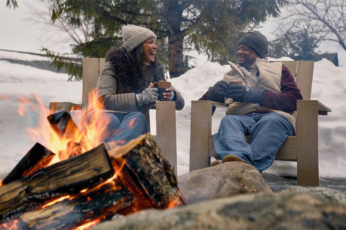 Two people sit in wooden chairs by a fire, dressed in winter clothing, holding mugs and smiling, with snow and pine trees in the background.