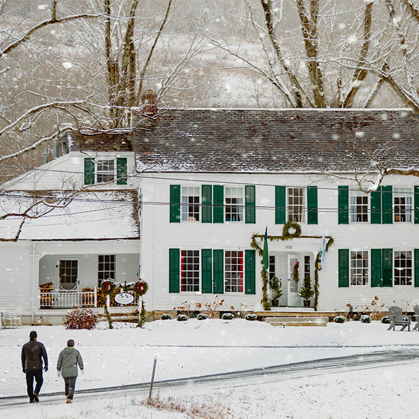 A white historic inn seen from outside in the winter on a snowy day. Two people walk toward it.