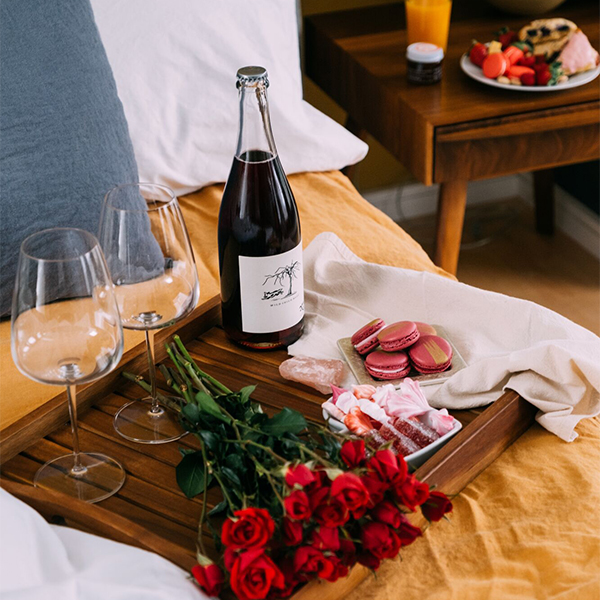 Romantic setup on a bed with wine, glasses, charcuterie board, and red roses.