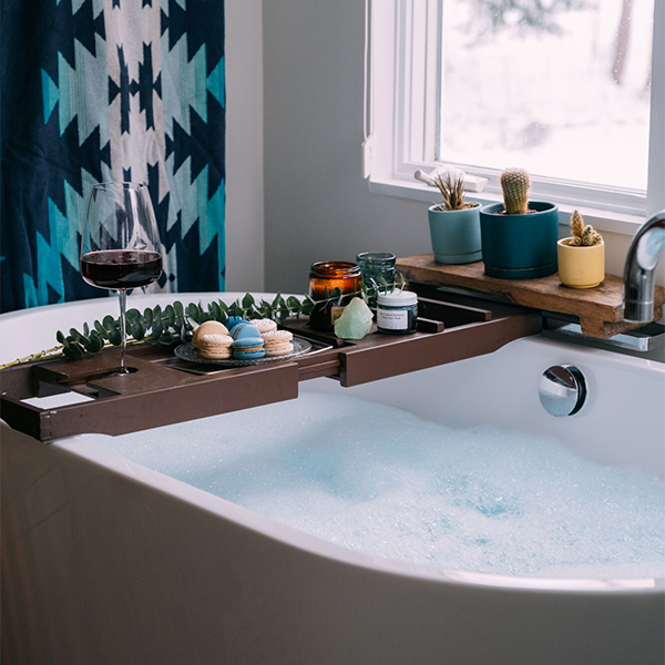 Bubble bath in a bathtub with a wooden bath tray holding wine, candles, and small plants.