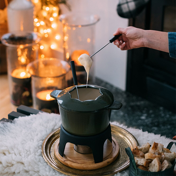 Person dipping bread into a pot of melted cheese fondue beside a cozy fireplace.
