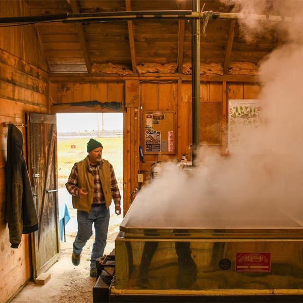 A person walks into a sugarhouse with steam visible from the boiler inside.