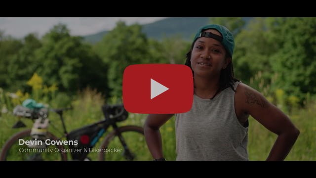 A video thumbnail featuring Devin Cowens, a community organizer and bikepacker. Devin, a person with a green baseball cap and grey tank top, stands in a green, grassy field with a loaded gravel bike behind her. A red YouTube play button is centered over the image.
