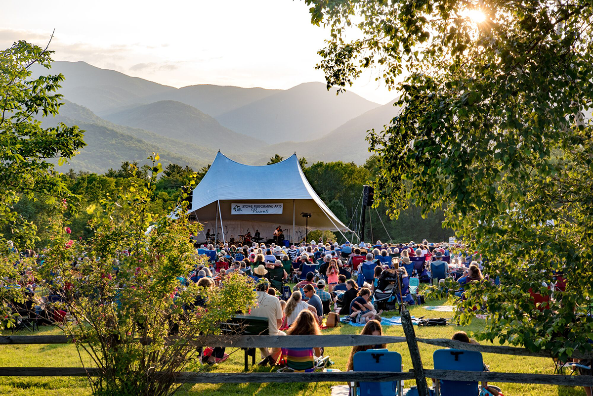 People gather around a tent to watch a live performance outdoors on a sunny summer evening.&nbsp;