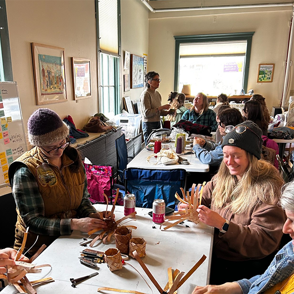 People gather inside a classroom-type space at tables smiling and working on an art project.  