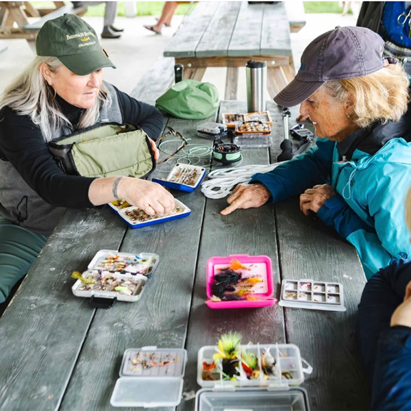 People sit at a wooden picnic table tying flies outside.&nbsp;