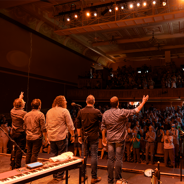 A band on stage seen from behind with people cheering in the background.&nbsp;