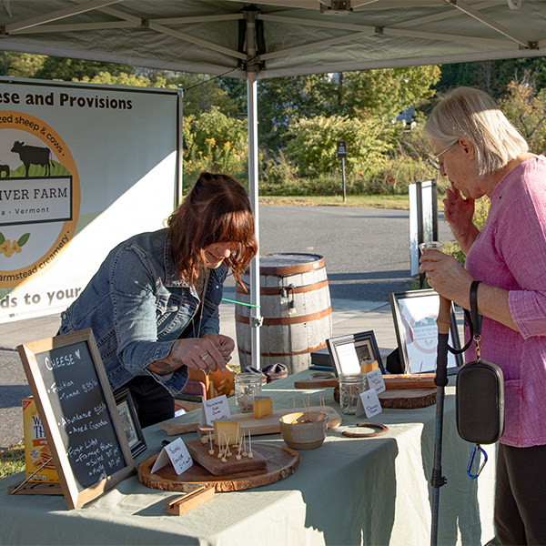 People sample cheese at a vendor table under a tent outdoors in the summer.&nbsp;