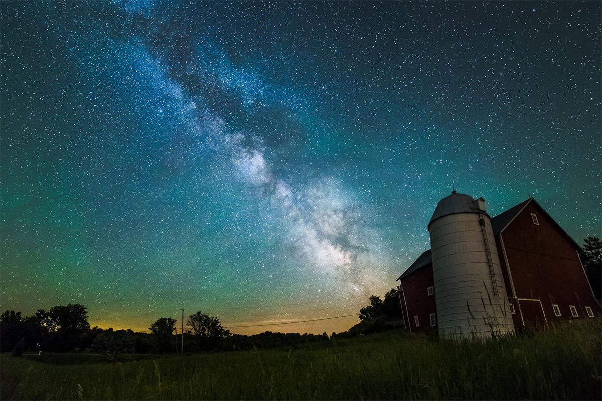 The Milky Way is prominently visible behind a silhouette of a barn at night.
