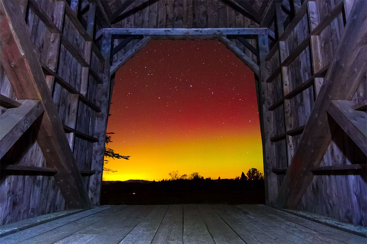 The aurora borealis, in bright red and yellow colors, visible through a wooden covered bridge at night. 