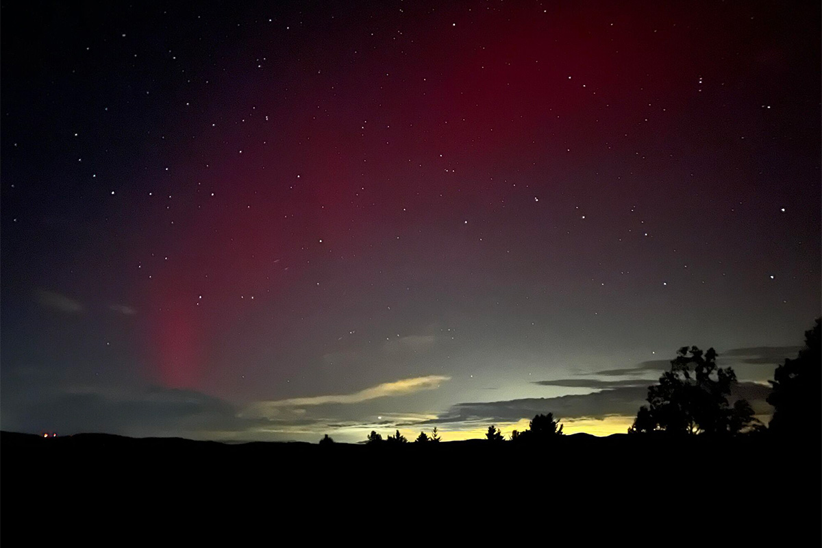 A deep red aurora borealis glows against a dark, starry night sky above a silhouetted treeline and horizon.