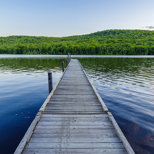 A dock stretches over a lake with a mountain covered in green trees visible on the other side. The sky is sunny and blue.&nbsp;