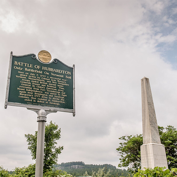 A green historical marker for the Battle of Hubbardton stands next to a tall stone obelisk monument under a cloudy sky.