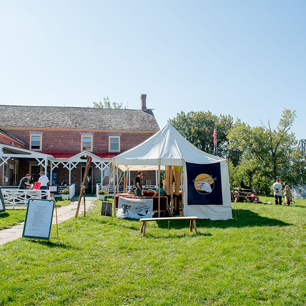 People gather outside a brick building at a historic site with a tent visible outside on a sunny summer day. 