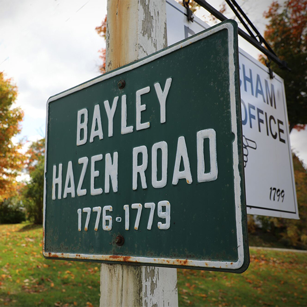 A sign reading “Bayley-Hazen Military Road, 1776-1779" outdoors in the late summer.