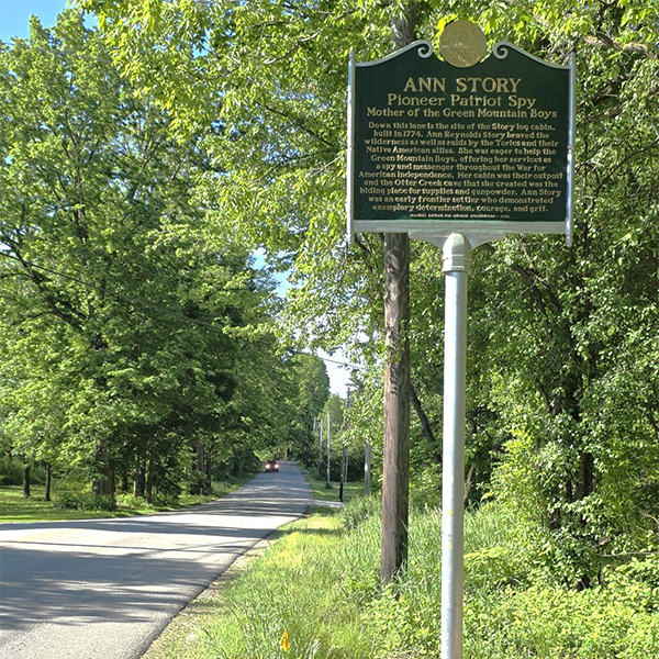 A historic marker sign next to a road reading “Ann Story: Pioneer, Patriot, Spy” seen on a sunny summer day.