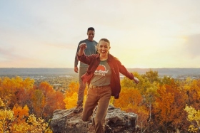 Two people walking near a cliff overlooking the fall foliage.