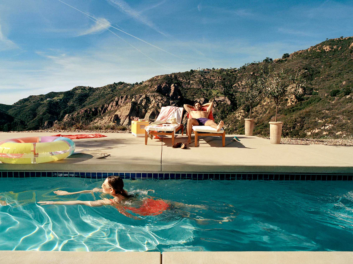 A woman swims in a pool while a man relaxes on a lounger with mountains in the background.