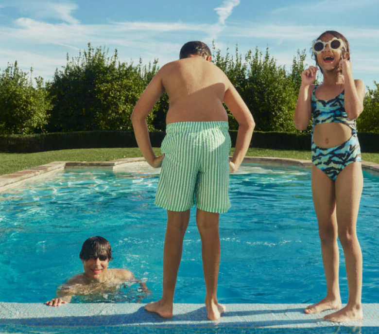 A girl in sunglasses looks at the camera while a boy contemplates the water poolside