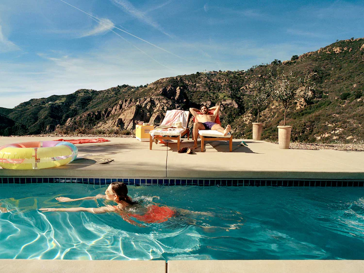 A woman swims in a pool while a man relaxes on a lounger with mountains in the background. 
