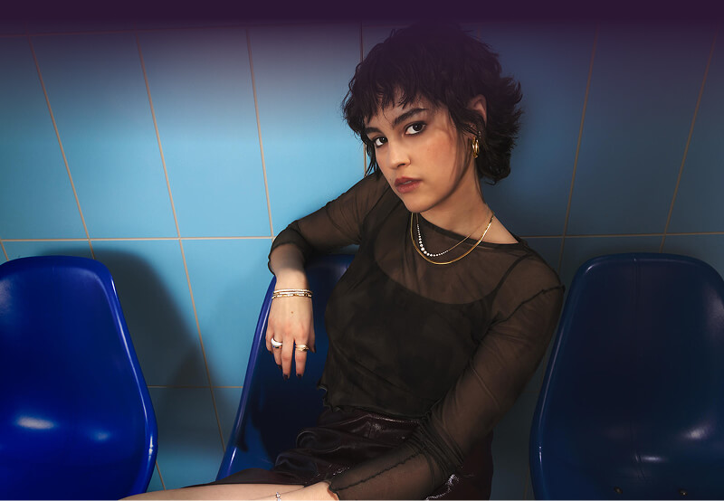 Woman in a sheer black top wearing stunning Necklaces, Bracelets, Rings and Earring sitting on a blue chair.