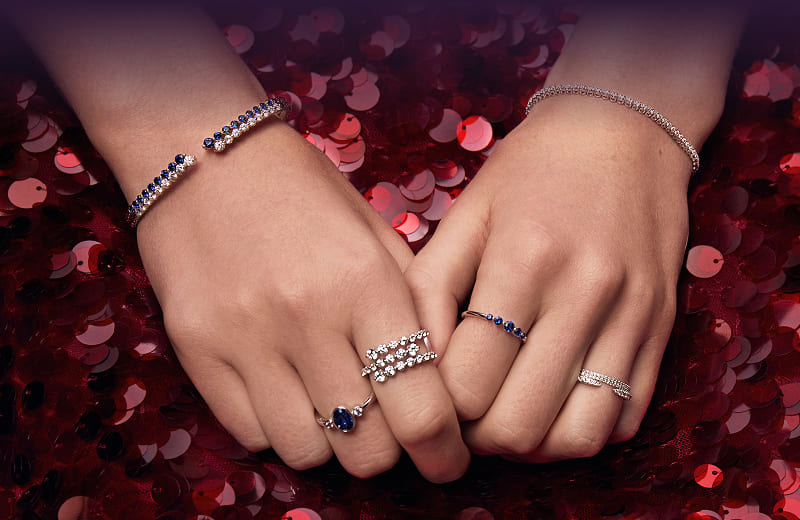 A model wearing gemstone rings and bracelets rests their hands on a red sequined surface.