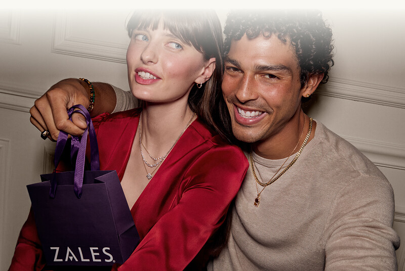 Male and female model smiling in diamond and gold jewelry, holding Zales shopping bag.