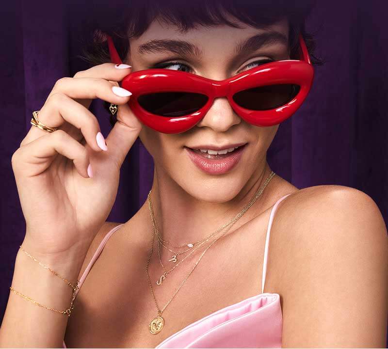 Model wearing bold red sunglasses, showcasing assortment of personalized gold jewelry with pendants, initials and pearls.