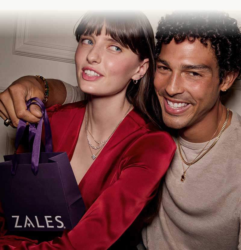 Female and male model, smiling wearing gold and diamond jewelry, holding Zales shopping bag.