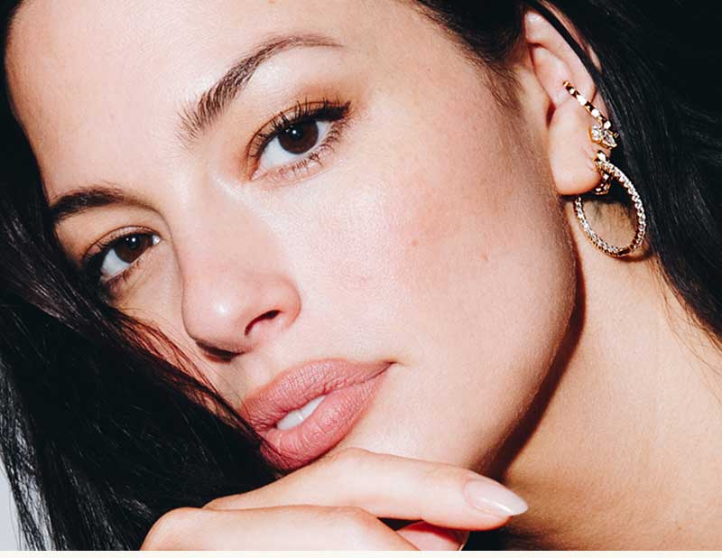 Close-up of Ashley Graham with gold ear cuff, gemstone studs & large hoops with diamonds. 