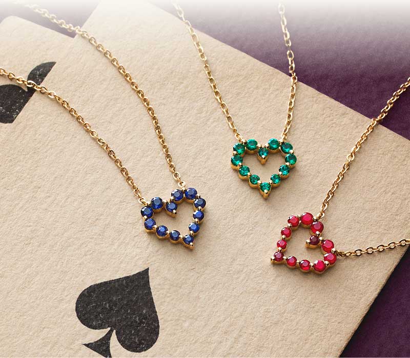 Gold chains with open-heart pendants with gemstones in blue, green & red displayed over a playing card.